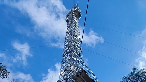 A shot looking up at the 140-foot tower with clouds floating above and tree leaves and branches seen in the bottom corners of the photo