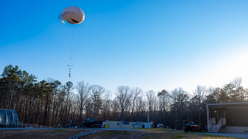 A tethered balloon system hovers over a forest clearing.
