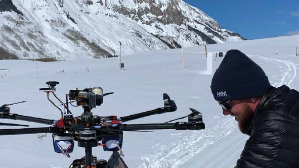 A person wearing a navy ARM beanie and black puffer jacket looks down at an open laptop screen next to a six-bladed uncrewed aerial system. In the distance are instruments spaced out across snow-covered ground and a large mountain.