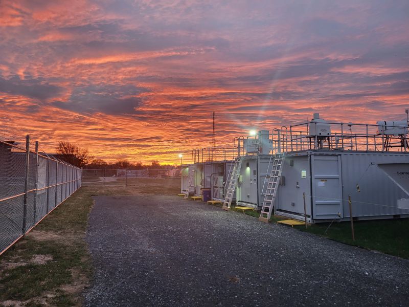 ARM instruments and containers line the edge of grass and gravel path. The sky is orange and cloudy. Street lights are seen in the distance.