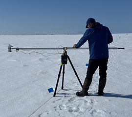 Standing on snow, Philip Wilson makes a broadband albedo measurement on Elson Lagoon during SALVO. The paired pyranometers are on the left end of the boom. Philip is focused on leveling the instrument using a bubble level directly in front of him on the boom.